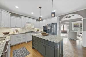 Spacious kitchen with white cabinets, granite countertops, and a large dark green island. Modern appliances and pendant lighting are visible. An open archway leads to a living area with a ceiling fan and sunlit windows. Hardwood floors throughout.
