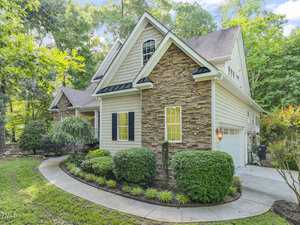 A charming two-story house with stone and siding exterior, surrounded by lush greenery. The home has a multi-gabled roof, shuttered windows, and a well-maintained front yard with shrubs and a curved concrete pathway leading to the entrance.
