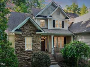 A charming two-story house with a stone and siding exterior, surrounded by lush greenery. It features a welcoming front porch and multiple windows. The roof is steeply pitched, and the sky is cloudy with hints of sunset colors.