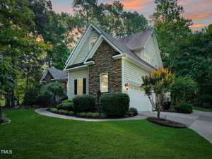 A charming house with stone accents and white siding is nestled among lush trees. It features a manicured lawn, bushes, and a double garage. The sky is a soft gradient of evening hues, adding warmth to the scene.