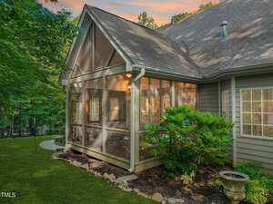 A screened porch with a peaked roof attached to a house, surrounded by greenery. Warm light glows from inside, and a pathway leads to the porch. The sky shows a colorful sunset.