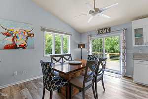 Dining room with a wooden table and chairs, large windows, and a sliding glass door leading to a deck. Walls are light blue with a colorful cow painting and a "HOME" sign above curtains. Ceiling fan overhead.