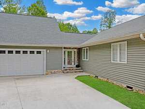 A modern gray house with a large garage and neatly trimmed lawn. The front entrance features a glass door with stone accents on the facade, under a clear blue sky with scattered clouds.