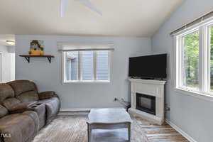 A cozy living room with a brown reclining sofa, a wooden coffee table, and a wall-mounted TV above an electric fireplace. The room has light blue walls, a high ceiling with a fan, and large windows letting in natural light.