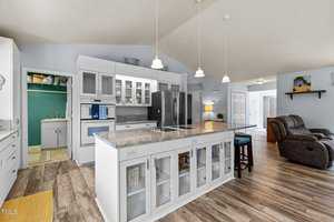 A modern kitchen with white cabinetry, an island with a marble countertop, and stainless steel refrigerator. Pendant lights hang above the island, and a cozy living area with recliners is visible in the background.
