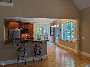 A kitchen and dining area with wooden flooring. The kitchen features dark wood cabinets, a bar with two stools, and stainless steel appliances. Large windows and glass doors provide natural light, and a chandelier hangs over the dining space.