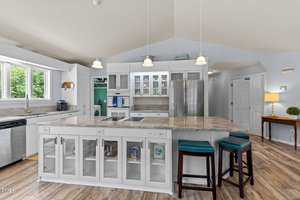 A spacious kitchen with white cabinets and a large island featuring glass-front storage and bar stools. Stainless steel appliances and pendant lighting add a modern touch. A window provides natural light, highlighting the wooden floor and light blue walls.