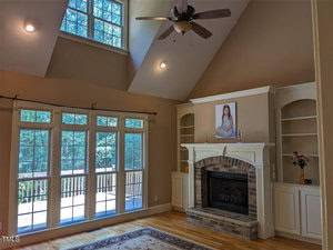 A spacious living room with high ceilings, large windows, and a ceiling fan. A brick fireplace is flanked by built-in shelves, and there's a portrait above the mantel. Sunlight streams in, illuminating the hardwood floors.