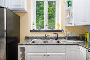 A kitchen scene featuring white cabinets, a stainless steel double sink, a black granite countertop, and a window showing green foliage outside. Various kitchen items are neatly placed, including a dish soap bottle and a banana on a stand.