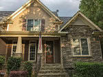 Nestled in Youngsville at 1004 Chatham Court, this cozy house boasts a mix of stone and wood siding. A front porch proudly displays an American flag, while the entrance is framed by neatly trimmed bushes under a partly cloudy sky.