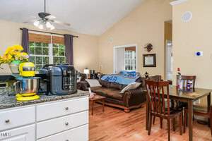 A cozy kitchen and living area featuring a granite countertop with a stand mixer and pressure cooker. A dining table with a flower vase sits nearby, while a sofa is visible in the background under large windows with natural light.