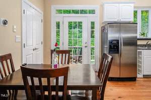 A kitchen with stainless steel appliances, a wooden dining table with four chairs, and a single rose in a vase. Large windows and glass doors in the background offer a view of greenery outside.