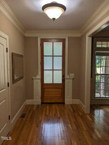A hallway with wooden flooring and beige walls, featuring a wooden framed door with glass panels. A mirror is hung on the left wall, while natural light from outside illuminates the space. A ceiling light fixture is overhead.