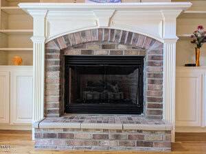 A traditional fireplace with a black metal firebox set within a red brick surround. The mantel, painted white with sculpted details, complements the built-in white cabinets and shelves on either side. A small vase with flowers decorates one shelf.