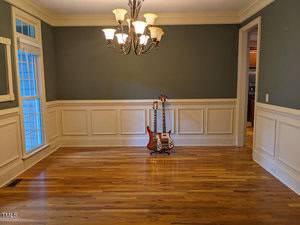 An empty room with green walls and wooden flooring features a chandelier and white wainscoting. A guitar rests against the wall near a window.
