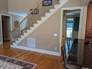 A wooden staircase with black metal railings ascends beside a door in a bright room. Hardwood floors and a patterned rug are visible. A kitchen island with bar stools is on the right, and sunlight streams through a window in the background.