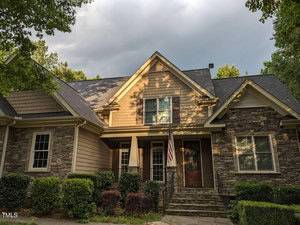 A charming house with stone and wood siding features a sloped roof and a covered porch. A small American flag is displayed near the entrance, and the front yard is landscaped with bushes and trees under a partly cloudy sky.