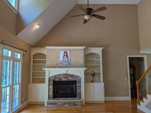 A spacious living room with a high ceiling features a brick fireplace flanked by built-in shelves and cabinets. Above the mantel is a portrait of a woman. The room is brightly lit by a large window to the left and a ceiling fan hangs from above.