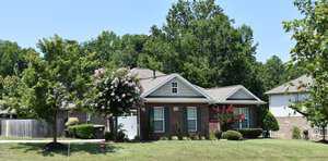 A brick house with a gabled roof and a well-maintained front yard. There are flowering crepe myrtle trees in front, surrounded by green grass and shrubs. Tall trees line the background under a clear blue sky.