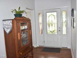 A hallway with a wooden cabinet displaying glassware and a plant on top. The entrance features a white door with decorative glass panels, allowing natural light to filter in. A dark mat is placed in front of the door on the wooden floor.