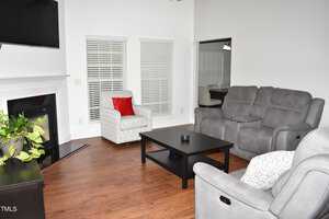 A cozy living room with a gray reclining sofa set, a patterned armchair with a red cushion, and a black coffee table. There is a fireplace on the left, wooden floors, and a potted plant. Large windows with blinds let in natural light.