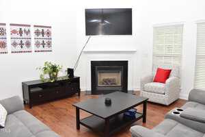 A modern living room with a mounted TV above a fireplace. There's a patterned chair with a red pillow, gray sofas, a black coffee table, a side table with a potted plant, and art on the walls. The floor is hardwood, and blinds cover the windows.