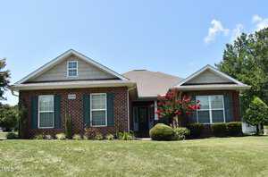 A single-story brick house with dark shutters features a well-maintained lawn and landscaping. Red flowering trees and bushes add color to the yard. The sky is clear and blue.