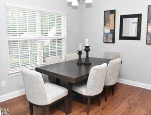 A dining room featuring a dark wooden table with six cushioned chairs. The table is topped with two decorative candle holders. The room has light walls, hardwood flooring, two abstract paintings, and a window with blinds.