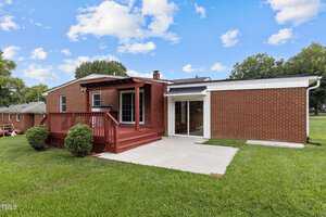 A single-story brick house with a red wooden deck in the backyard. The yard is grassy and well-maintained, with a few bushes near the deck. The sky is partly cloudy and clear.