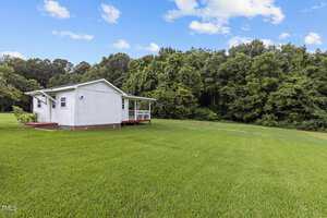 A small white house with a porch sits at the edge of a large, lush green lawn, bordered by dense trees under a partly cloudy blue sky.