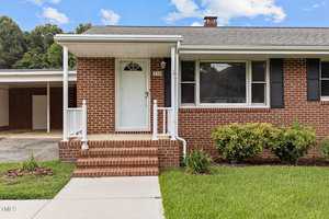 A brick house with a white front door and steps leading up to it. There is a small porch with a white railing. The house has black shutters and is surrounded by a green lawn. A carport is visible in the background under a partly cloudy sky.