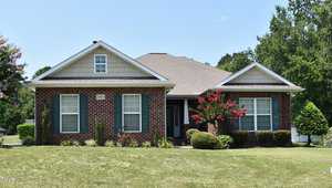 This charming single-story brick house on 2005 Massimo Drive in Creedmoor boasts a gabled roof, white trim, and black shutters. It's surrounded by a well-maintained lawn, small trees, and bushes. A bright red flowering tree stands near the entrance against a backdrop of clear blue skies.