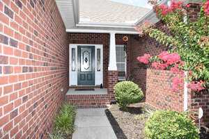 A brick exterior entrance with a dark green front door framed by white sidelights. Neatly trimmed bushes and flowering plants line the walkway leading to the porch. A small window is on the right of the door.