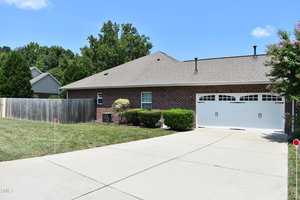 A brick house with a sloped roof and a two-car garage is situated on a concrete driveway. The yard is well-kept with green bushes. A wooden fence is visible on the left, and there are trees in the background under a clear blue sky.