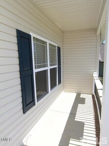 A narrow porch with beige siding and a white railing, featuring a window with dark blue shutters. The porch casts striped shadows from the sunlight filtering through the railing.