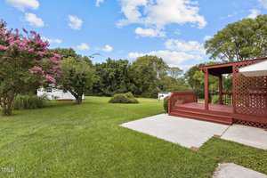 A backyard with a wooden deck and lattice, surrounded by a lush green lawn. A flowering tree and bushes are on the left, and two white sheds are visible in the distance under a blue sky with clouds.