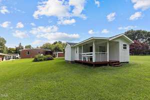 A small white cottage with a porch is set on a grassy lawn under a blue sky with scattered clouds. A larger red brick building is visible in the background, surrounded by trees and bushes.