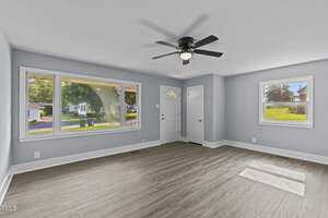 A bright living room with light gray walls and wood floor features a ceiling fan, large windows, and a white front door. Sunlight streams in, and the view outside shows greenery and neighboring houses.