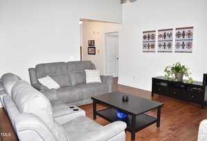 A living room with gray sofas, a black coffee table, and a TV stand. There are patterned artworks on the white wall and a plant on the TV stand. The floor is wooden, and light enters from the left.
