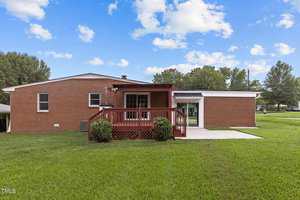A brick house with a red wooden deck is set against a lush green lawn under a partly cloudy sky. Small shrubs line the edge of the deck, and a concrete pathway is visible on the side of the house, leading to a glass door.