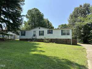 Nestled at 200 Pecan Street in Creedmoor, a white single-story house with blue shutters and a brick foundation sits surrounded by trees on a sunny day. The yard is grassy, and a small set of stairs leads to the welcoming front door.