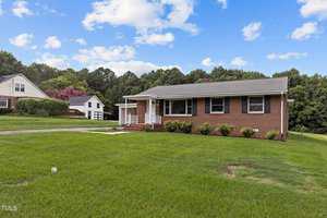 A one-story brick house with a gray roof is situated on a lush green lawn. The yard is bordered by trees, and a garage is visible in the background. A driveway leads to the front steps, and the sky is partly cloudy.