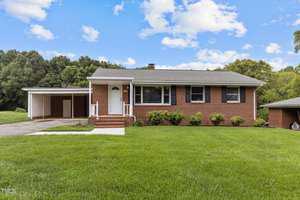 Nestled at 210 Grove Street, this charming brick house in Oxford features a large front yard, a convenient carport, and steps leading up to the welcoming front door. Surrounded by lush trees under a partly cloudy sky, it offers both tranquility and curb appeal.