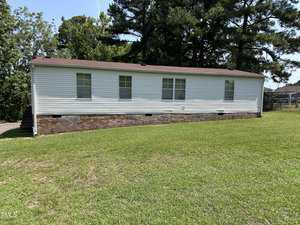 A white single-story mobile home with a red-brown shingled roof sits on a grassy lawn. The house has several windows and brick skirting, surrounded by trees under a clear blue sky.