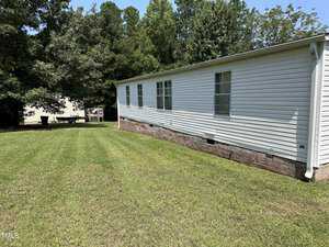 A single-story white mobile home with several windows is situated on a grassy lawn. In the background, there are dense trees and a small structure partially visible. The sky is clear and sunny.