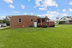 A brick house with a sloped red roof and wooden deck is surrounded by a well-manicured lawn. The sky is blue with scattered clouds, and neighboring houses are visible in the background.