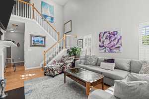 A bright living room featuring a gray sectional sofa with patterned pillows, a floral armchair, and a wooden coffee table. Artworks adorn the light gray walls, and a staircase with wooden railings is visible in the background. Natural light fills the room.