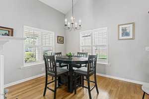 A bright dining area features a dark wooden table with four matching chairs. Large windows with white shutters let in natural light. A modern chandelier hangs above the table, and framed art decorates the light gray walls.