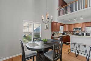 A modern kitchen and dining area with high ceilings. There's a dark wooden dining table with six chairs and a bowl of green apples. The kitchen features brown cabinets, stainless steel appliances, and a small bar area with stools. A chandelier hangs overhead.
