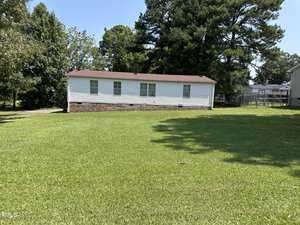 A small white mobile home with a red-brown roof sits on a large, neatly trimmed grassy lawn. Tall green trees surround the house, and a fence is visible on the right side.